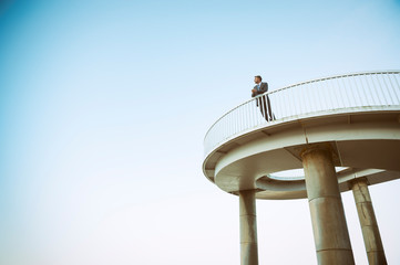 Distant businessman standing alone on curved elevated walkway under soft blue sky