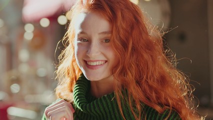 Portrait of cute young woman with curly red hair in a bright sunlight looking and smiling at camera walking in the city