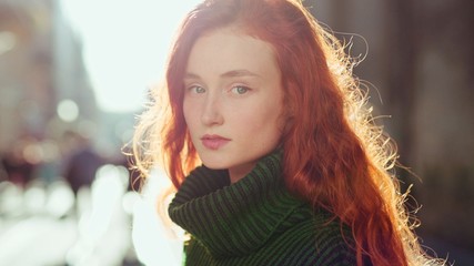 Portrait of charming young woman with red hair in green sweater smiling at camera and posing in the city sun shining brightly feel happy having fun