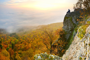 Alone Man sitting on the top of the rock under the mist, sunrise sky and beautiful colorful autumn nature