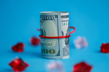 A roll of dollars wrapped in a red rope ribbon on a background of scattered red and pink crystals, a 100 dollar