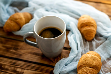 on a wooden table with blue gauze is a gray cup with coffee nearby in blur croissants
