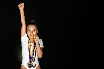 Portrait of smiling little gymnast girl in white bodysuit with medals on her neck biting a medal,...