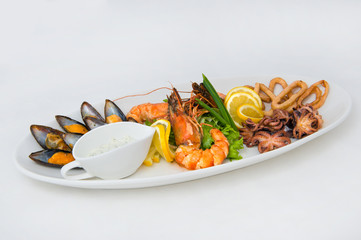 On a long oval plate an abundance of seafood. Mussels, king prawns, small octopuses, squid rings. As well as white sauce with herbs, lettuce, lemon, onion feathers.