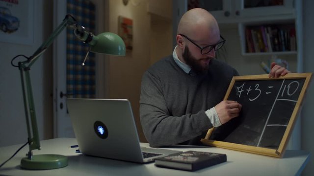 Bearded 30s Male Teacher In Glasses Writing Math On Chalk Board With White Chalk Looking At Laptop At Home. Online Education Process. Side View Of Teacher Talking. 