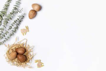 Wooden Easter eggs in a straw nest, decorated by wooden Easter bunnies and dried Eucalyptus leaves, on a white background.