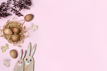 Wooden Easter eggs in a straw nest, decorated by mini wooden Easter bunnies, larger Easter bunny figures and dried Ruscus leaves on a pink background.