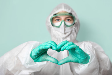 Medical worker in safety glasses, mask and suit makes a heart sign. Green background. Confronting...