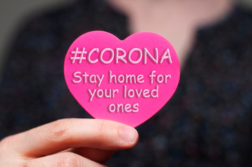 Caucasian woman holding pink heart shaped sticky note or card. Text saying Corona stay home for your loved ones. Coronavirus quarantine concept, social distancing concept. Stop virus from spreading.