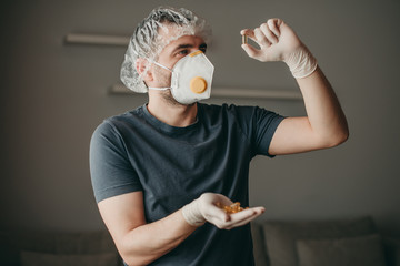 brunette white european man doctor with medical mask on gray background holding a pill of vitamin omega 3. Coronavirus, illness, infection, quarantine, surgical bandage, respirator with valve.