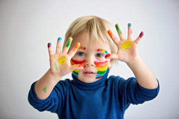 Beautiful blond toddler boy with rainbow painted on his face and messy hands