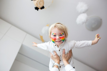 Beautiful blond toddler boy with rainbow painted on his face and messy hands