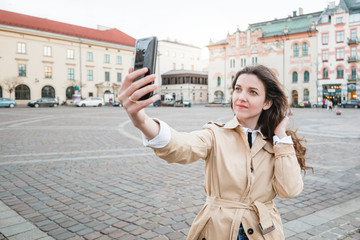 Fototapeta premium Happy young woman taking selfie at city street. Happy tourist girl walking outdoors. Spring fashion portrait of pretty brunette female posing in old town.