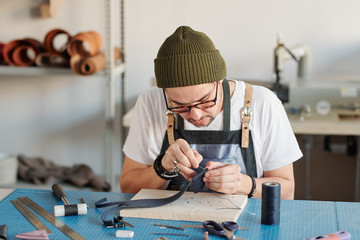 Young creative leatherworker bending over table while sewing new item