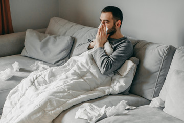 Young sick bearded european man sneezes into napkin at home on gray sofa with white blanket....