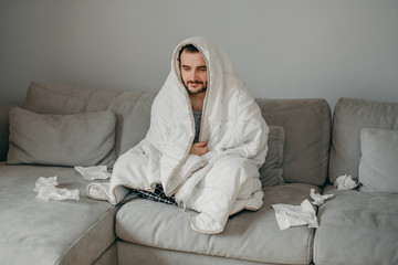 Young sick bearded european man sneezes into napkin at home on gray sofa with white blanket. Disease, protection, coronavirus, virus, disease, flu, respiratory dressing.