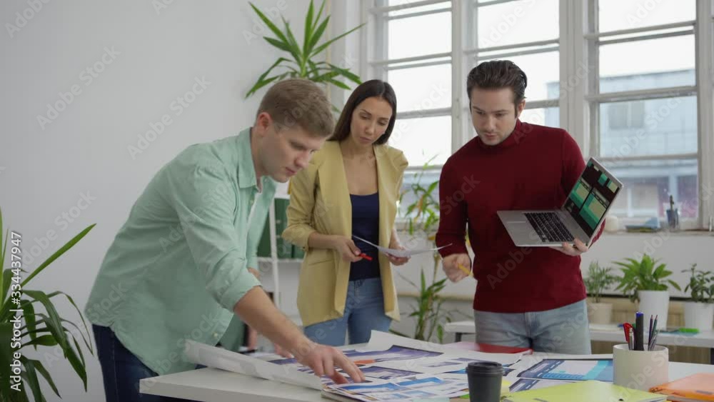 Wall mural tracking medium shot of young startup team of three specialists standing around table in office and 