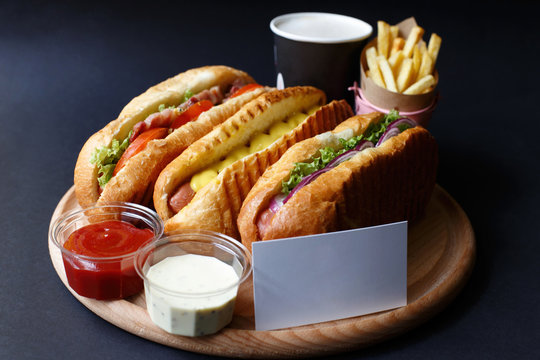 Three hot dogs, sause, ketchup, coffee, French fries lying on the wooden plate.