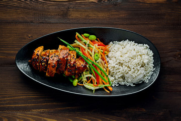 Delicious Asian teriyaki chicken, salad made of cucumber, carrot, ginger, pepper and cabbage, edamame, sesame and rice in a black pottery plate. Top view, directly above.