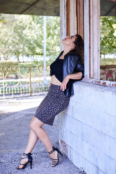 Young brunette woman, wearing black top with leather jacket and dark long skirt and high heels, posing in old abandoned building. Urban lifestyle female photo session.