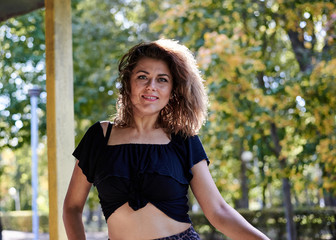 Young brunette woman, wearing black top, leather jacket and dark long skirt and high heels, posing near yellow rusty fence in city park. Urban lifestyle female photo session in summer autumn spring.