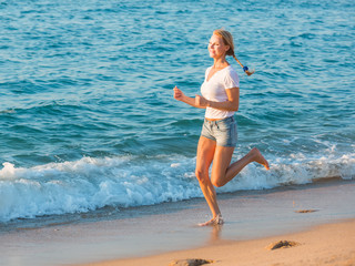 Smiling adult woman in white T-shirt is jogging on the beach