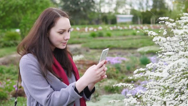 Happy long-haired girl makes a photo of blooming flowers on the smartphone in spring