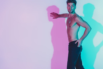 sexy shirtless man in dark blue jeans touching wall, holding hand in pocket and looking away on background with blue and violet shadows