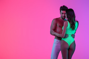full length view of sexy man in white jeans embracing seductive girl in green swimsuit on pink and purple background
