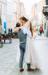 Happy groom holds his bride in hands kissing on lips against morning street background