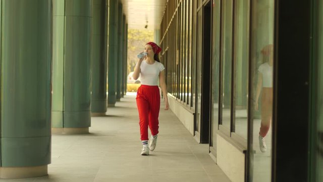 Girl drinks water from plastic bottle teenager kid walks street sidewalk in red pants outdoors