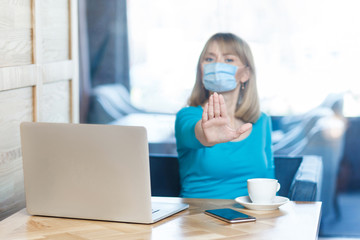 Stop, don't come any closer. Portrait of serious young woman with surgical medical mask sitting and showing stop with hands and warning about distance. Indoor working, medicine and health care concept