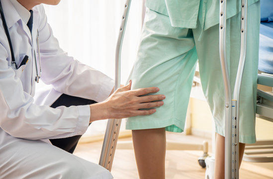 Asian male Doctor or Physiotherapist examining knee and helping patient to walk with crutches because injured legs of young patient women after accident and explain to her the diagnosis of problem.