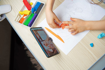 Little girl using smartphone to have online lesson a plasticine drawing lesson. Online education concept. 