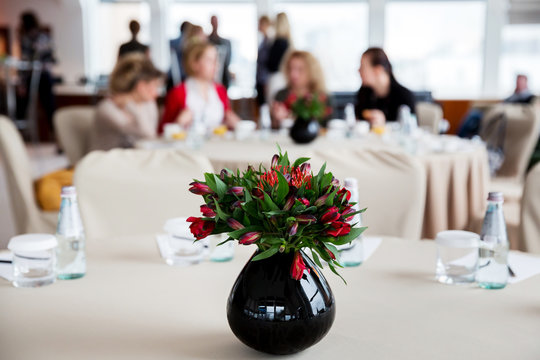 Bouquet of beautiful flowers in jug standing on a large round table for decoration. Business women at conference sitting in the background on the negotiations at conference room