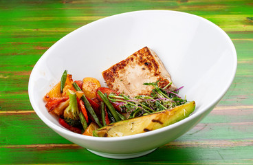 Fried tofu with grilled avocado and wok vegetables. On a colored wooden background. Vegetarian dish.