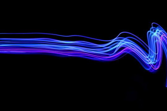 Long exposure photograph of neon blue and purple streaks of light in an abstract swirl, parallel lines pattern against a black background. Light painting photography.