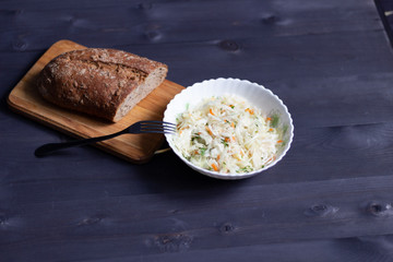 Fresh cabbage salad with carrot and dill in white bowl and brown bread on black wooden background