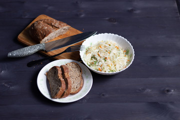Fresh cabbage salad with carrot and dill in white bowl and brown bread slices and foaf on black wooden background