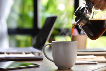 A hand pouring steaming coffee in to a cup on a work desk when work from home - 334462355