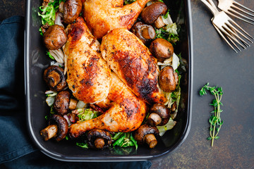 Top view of roasted chicken legs and breasts with fresh salad and mushrooms in black dish.