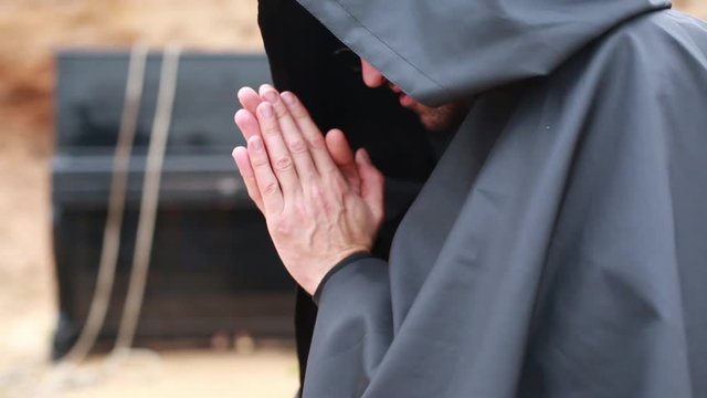 A monk in a black robe with a hood sits on a vacant lot and warms himself by the fire