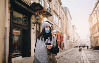 Fototapeta na wymiar A young girl in a hat, a coat of a protective blue medical mask-respirator with a smartphone in her hand walks along the evening street of a deserted city. Quarantine, coronavirus, pandemic COVID-19.