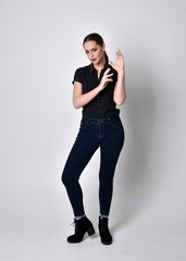 Portrait of a pretty brunette girl with ponytail, wearing jeans boots and a black blouse.  full length standing pose on a studio background.