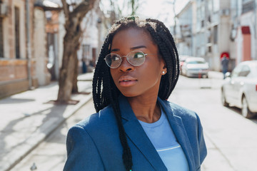 Close up portrait of a beautiful young african american woman with pigtail hairstyle in a blue jacket and glasses smiling and walking along the street