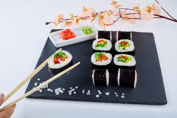 Sushi set with ginger and wassabi on a black square plate of stone on a light background. Near a sprig of sakura, a female hand holds sushi with chopsticks.