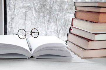 Round reading glasses lie on an open book with white pages next to a stack of multi-colored books on a windowsill
