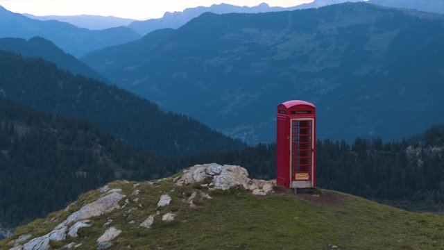 Aerial view of the red telephone box as a modern art object on the green field on the top of the mountain in the French Alps with a lake in the background and other mountain peaks.