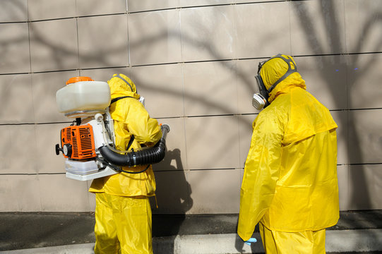 Paramedics Wearing Yellow Protective Costumes And Masks Disinfecting Coronavirus With Motorized Backpack Atomizer And Sprayer