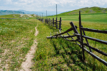 Broken wood fence and poles with wires along vast field in mountains in sunny day. Beautiful sunny alpine landscape with asphalt road along field behind long fence in highlands. Vivid mountain scenery